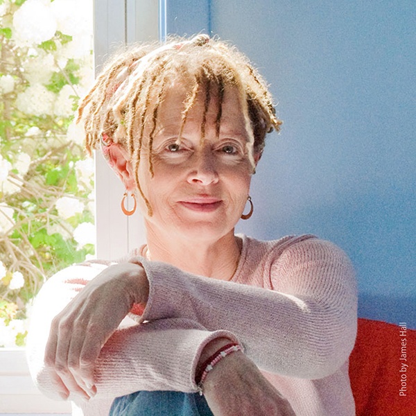 Anne Lamott sitting on a red chair in a light knit sweater, arms cross on her knee. The wall is blue behind her with a window full of a bush in bloom.