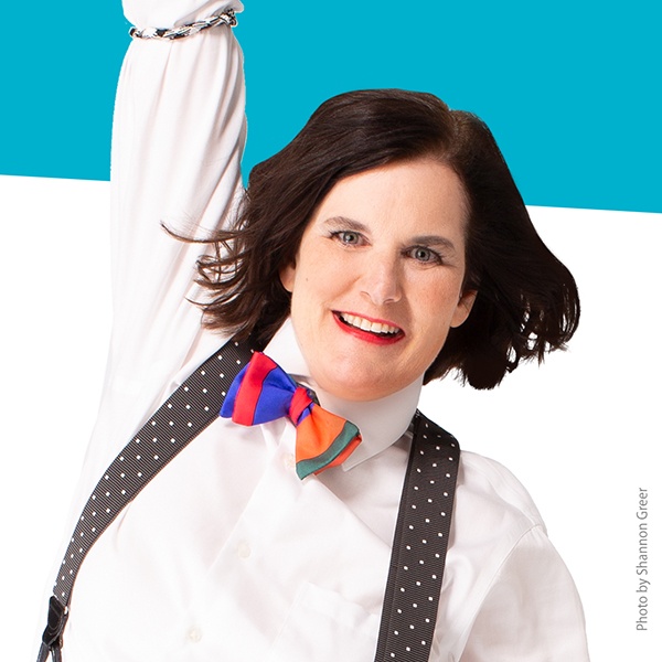 Paula Poundstone smiling with one arm raised wearing white-dotted suspenders and a colorful bow tie.