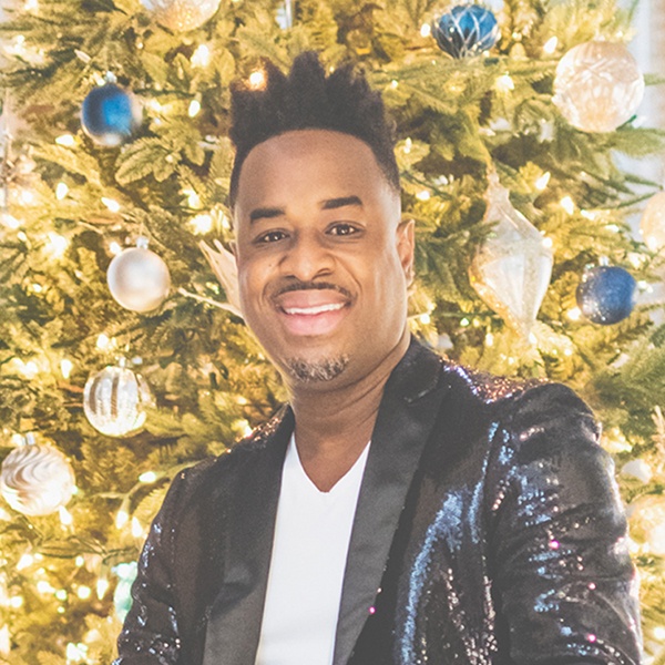 Damien Sneed in a sparkling dark jacket in front of a brightly decorated Christmas tree.