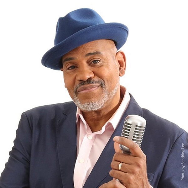 Allan Harris in a blue hat and suit, holding a microphone and smiling to the camera.