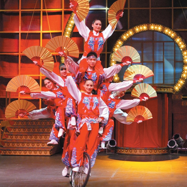 Members of the troupe holding fans and balancing on a single bicycle