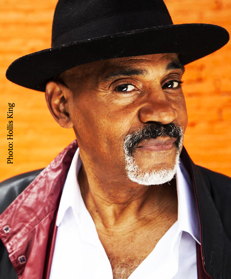 Allan Harris standing in front of a brick wall, dress in a jacket and hat.