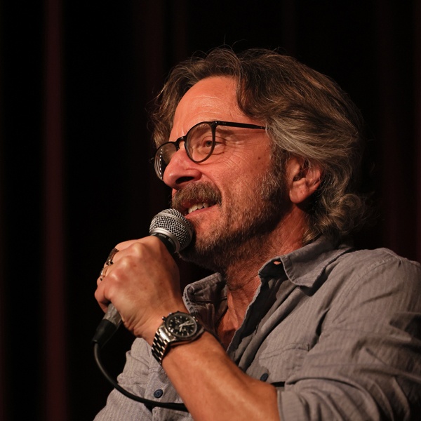 Marc Maron on stage, holding a microphone and looking at the
