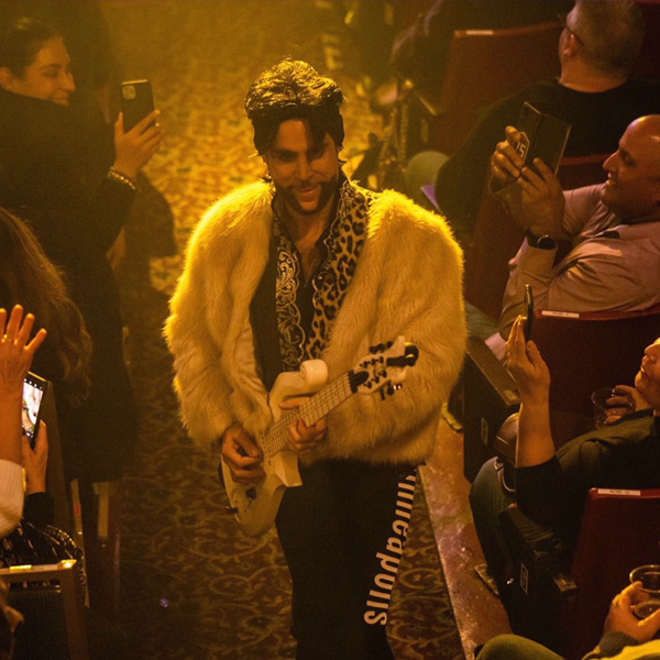 Marshall Charloff as Prince in a fur coat playing guitar in the aisle of a venue while people look on filming him on their phones.