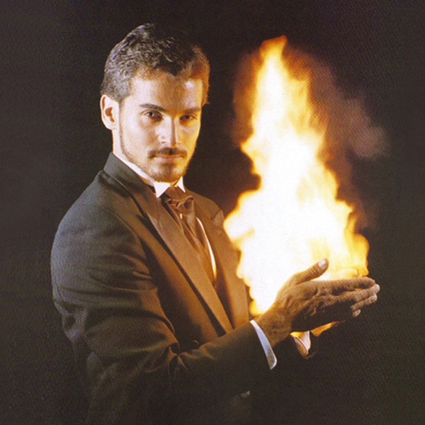 Dan Birch in a suit and tie staring mesmerizingly into the camera while holding a flame in his hands.