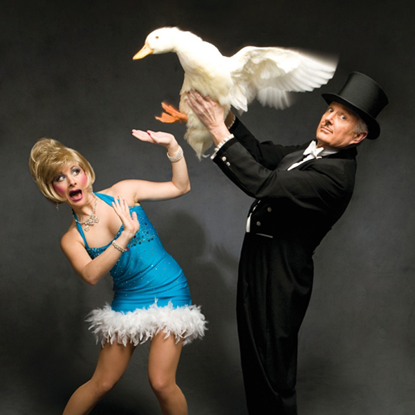 Les Arnold in top hair holding a goose while his assistant Dazzle, in a blue dress, cowers.