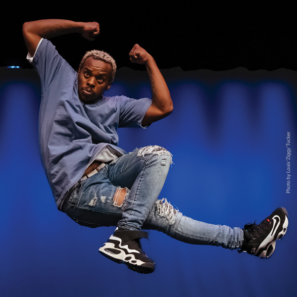 Lil Buck in the air, arms overhead, one leg crossed over the other.