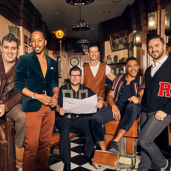 Members of The Doo Wop Project sitting in an old-fashioned barber shop smiling at the viewer.