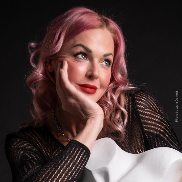 Storm Large in a striped black top and pink hair, holds her chin in her hand and looks off into the distance.