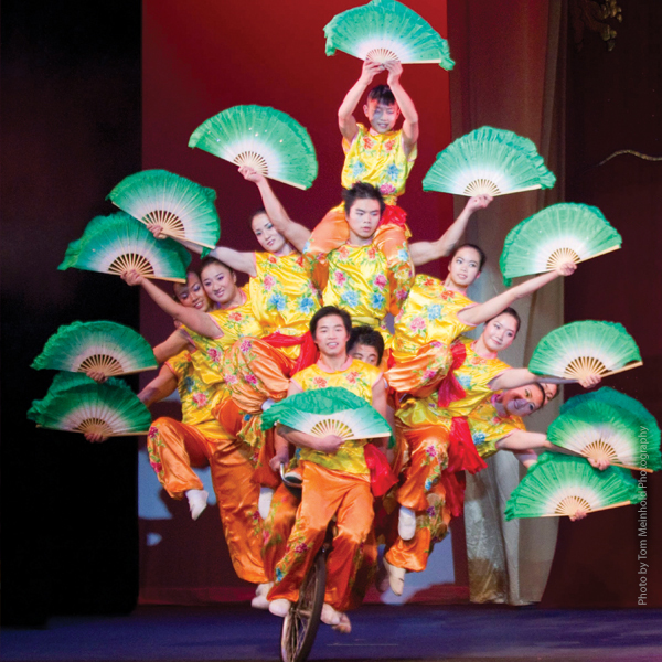 Members of The Peking Acrobats balancing together on a bicycle, jade-colored fans held aloft.
