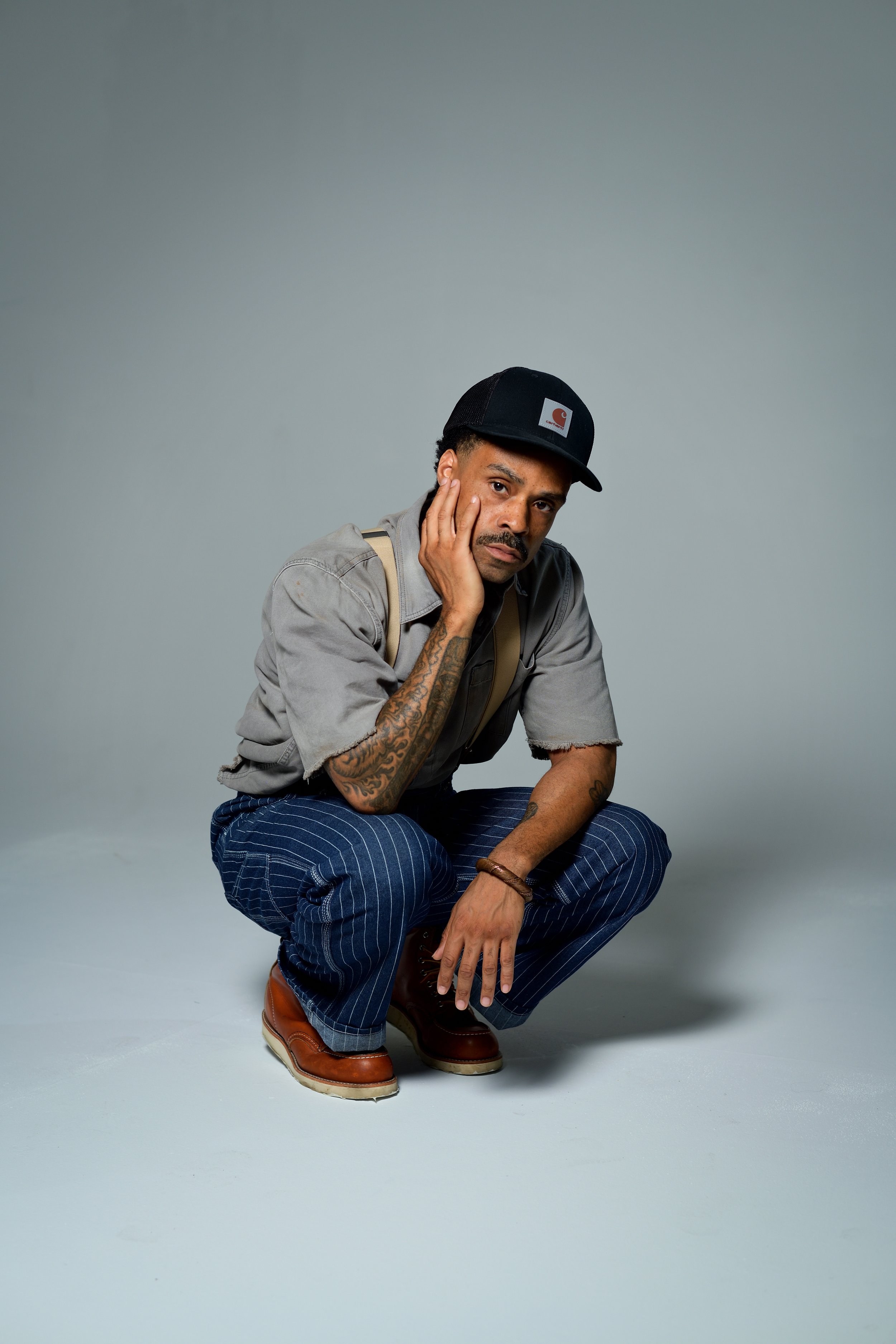 Bilal in a hat crouched with one hand against his cheek.