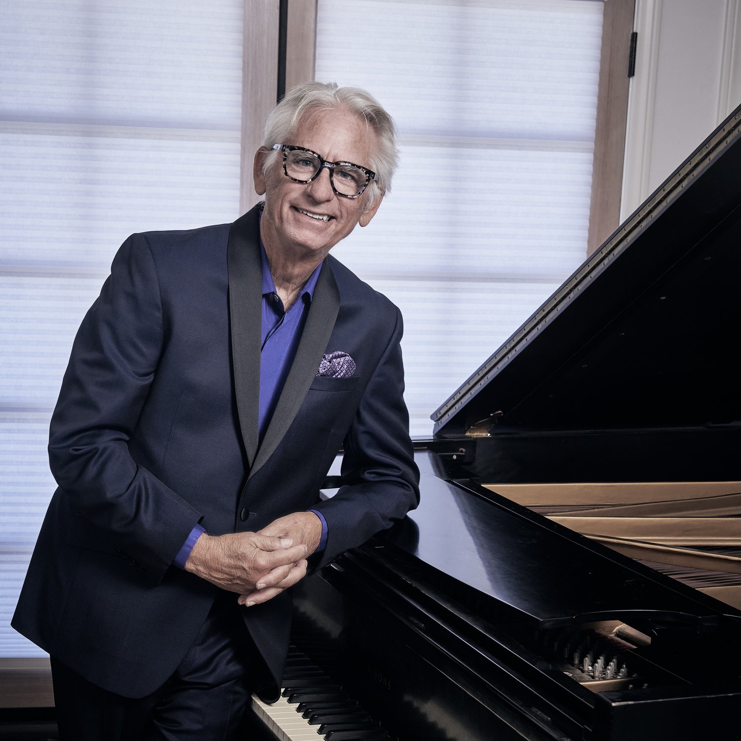 David Benoit in a dark suit leaning against a grand piano.