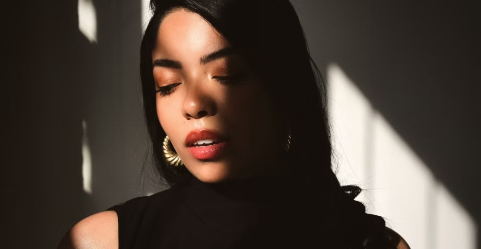 Portrait of a Lucía with long dark hair, wearing gold hoop earrings and a black top, bathed in warm sunlight with dramatic shadows cast across her face and background. Her eyes are closed, creating a serene and contemplative mood.