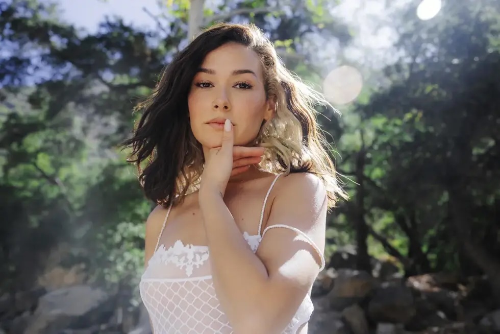 Nella in a white lace top poses outdoors, holding one finger to her lips.