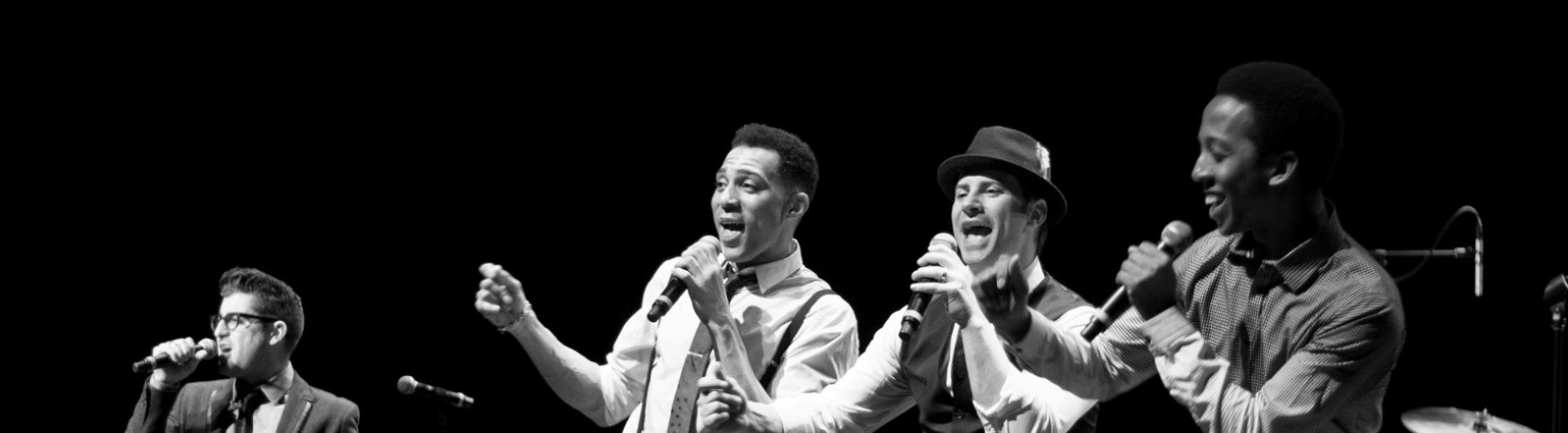 Members of The Doo Wop Project singing on stage against a dark background.