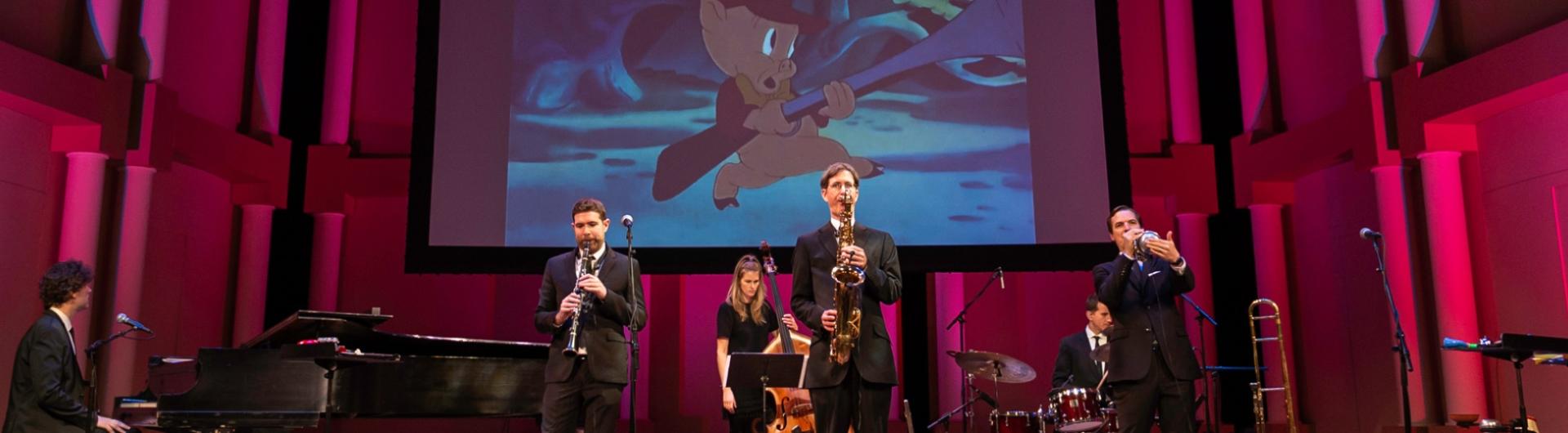 Members of the band performing on stage while a classic cartoon plays on a screen behind them.