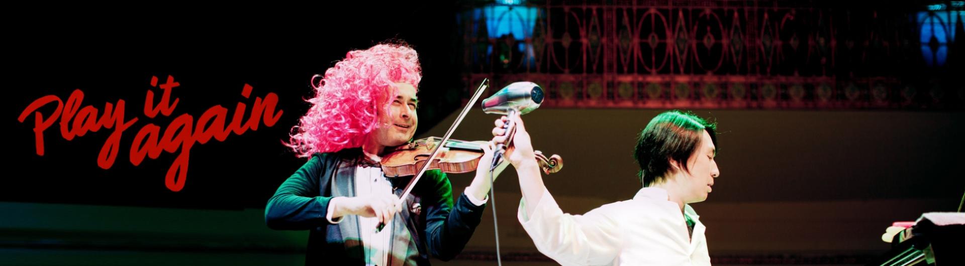 Igudesman dressed in a curly pink wig is playing the violin while Joo, at the piano, holds a hair dryer at him.