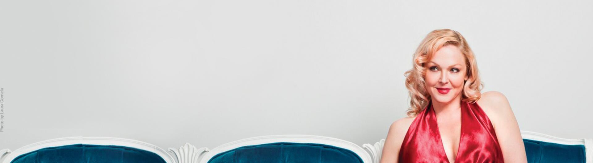 Storm Large in a red dress sitting on a chair and smiling.