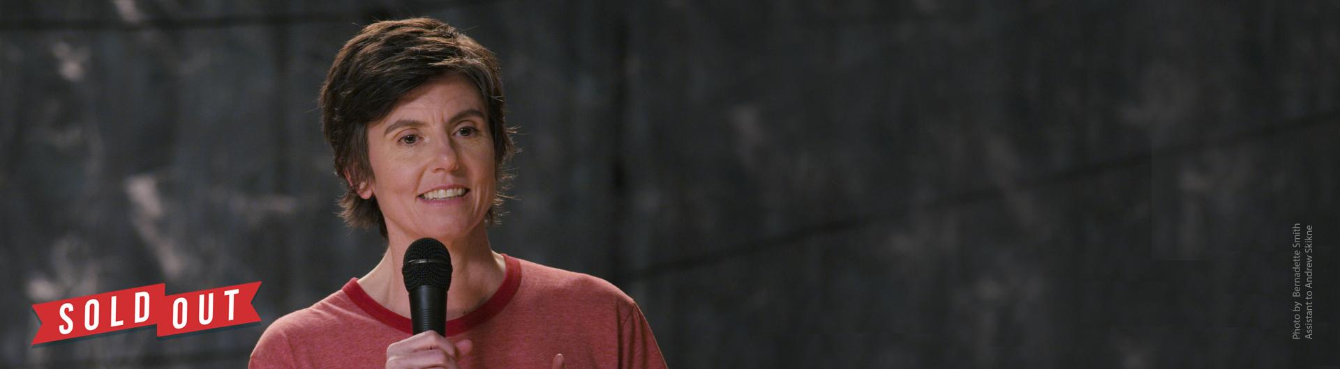 Tig Notaro on stage in a red T-shirt holding a microphone. The text SOLD OUT, referring to tickets, is displayed in the corner on a ribbon.