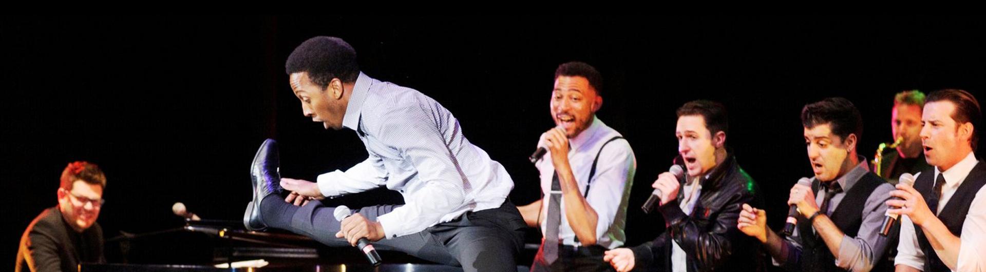 Members of The Doo Wop Project performing on stage holding microphones while one member does the splits and another plays the piano.