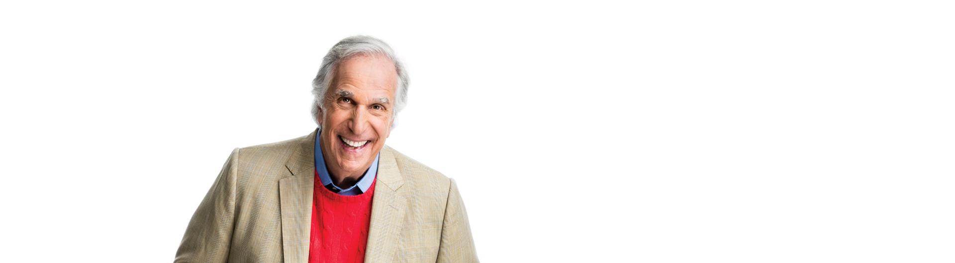 Henry Winkler in a red sweater and tan jacket smiling at the viewer.