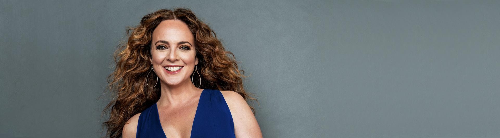 Melissa Errico in a dark blue dress smiling at the camera against a gray background.