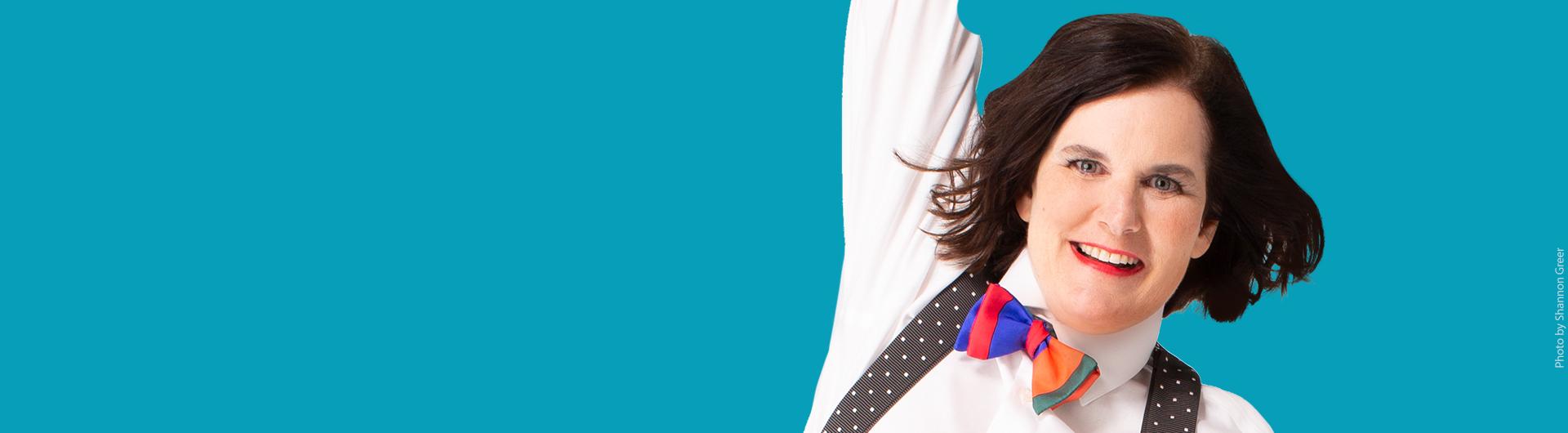 Paula Poundstone in a white shirt, suspenders, and colorful bow tie with one arm reaching upwards.