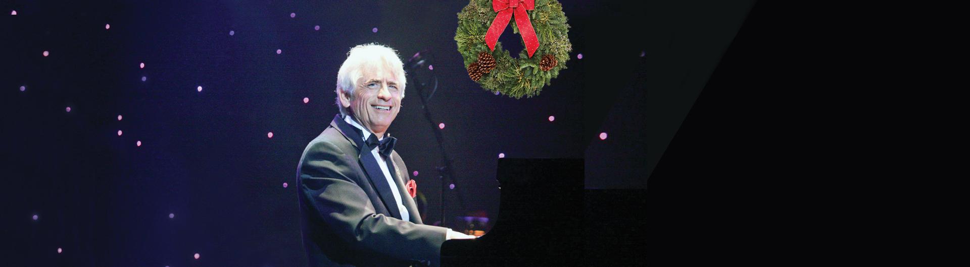 David Benoit sits smiling at a grand piano, the background filled with pinpricks for stars, a holiday wreath overhead.