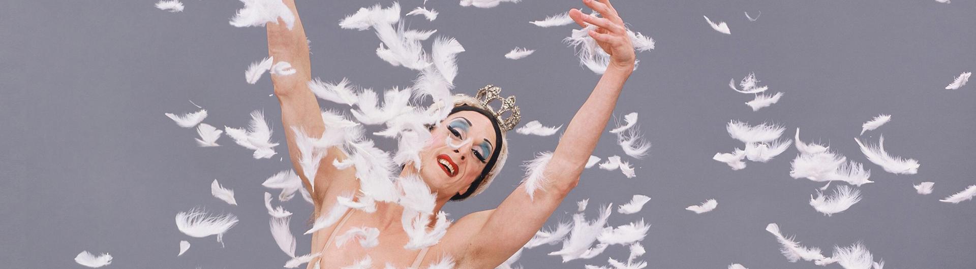 A member of the Trocks, arms outstretched, white feathers drifting to the ground.