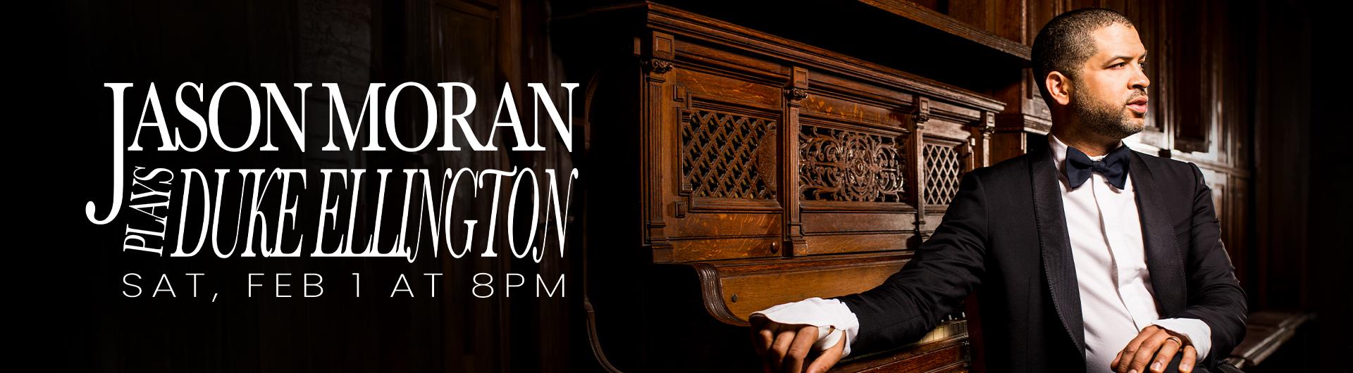 Jason Moran in a black jacket and bow tie sitting with his back to an upright piano.