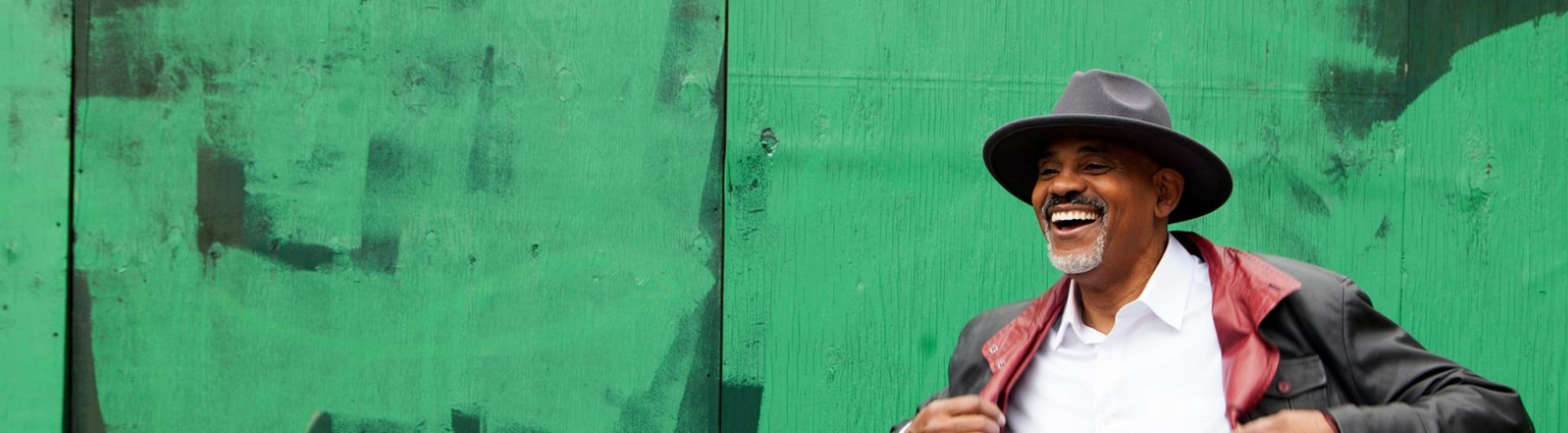 Rev Alt Text Allan Harris in a white shirt, hat, and jacket in front of an abstract green background.