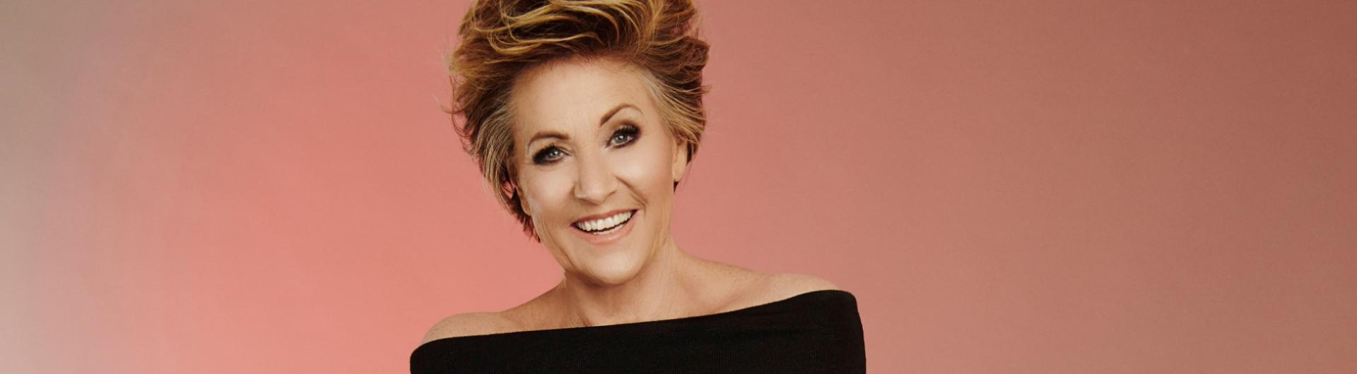 Lorna Luft in a black top, bare shoulders against a peach colored background.