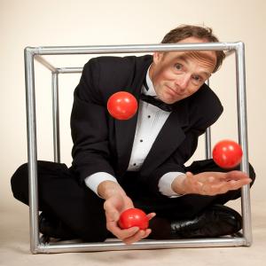 Henrik Bothe in formal attire, crouched within a cube while juggling red balls.