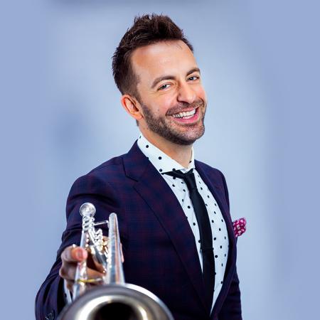 Benny Benack holding a trumpet toward the camera and smiling in a dark blue suit and tie