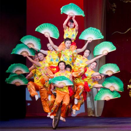 A dozen members of The Peking Acrobats holding green fans and balanced on a bicycle.