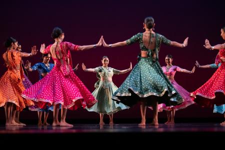 Sa Dance Company performers in colorful traditional dress dance in formation on a darkened stage.