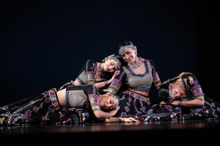 SA Dance Company performers in ornate black costumes form a reclining tableau on a dark stage.
