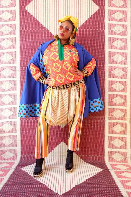 Portrait of Mumu Fresh in colorful patterned clothing and sunglasses, standing against a geometric textile backdrop.