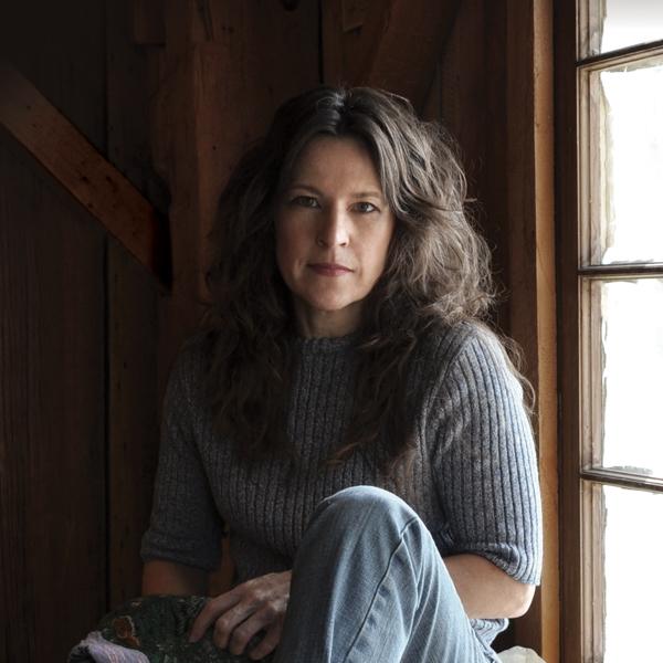 Carole O King in a sweater near a window in a photo that pays homage to the Carole King album cover for Tapestry.