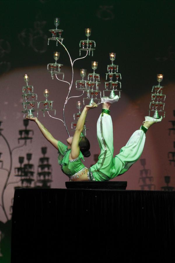 A female acrobat dressed in a green costume performs an intricate balancing act on a black platform, holding multiple ornate candelabras with her hands, feet, and mouth. Each candelabra is stacked with delicate glass tiers, some with lit candles, as she poses in a dramatic backbend under theatrical lighting.
