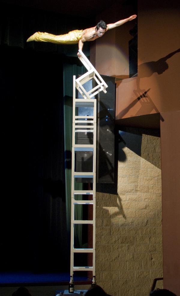 A male acrobat balances horizontally on one arm atop a precarious stack of white chairs, arranged in a vertical tower on stage. He wears gold pants and extends his free arm outward for balance. The dramatic lighting casts shadows against the theater wall, emphasizing the height and tension of the stunt.