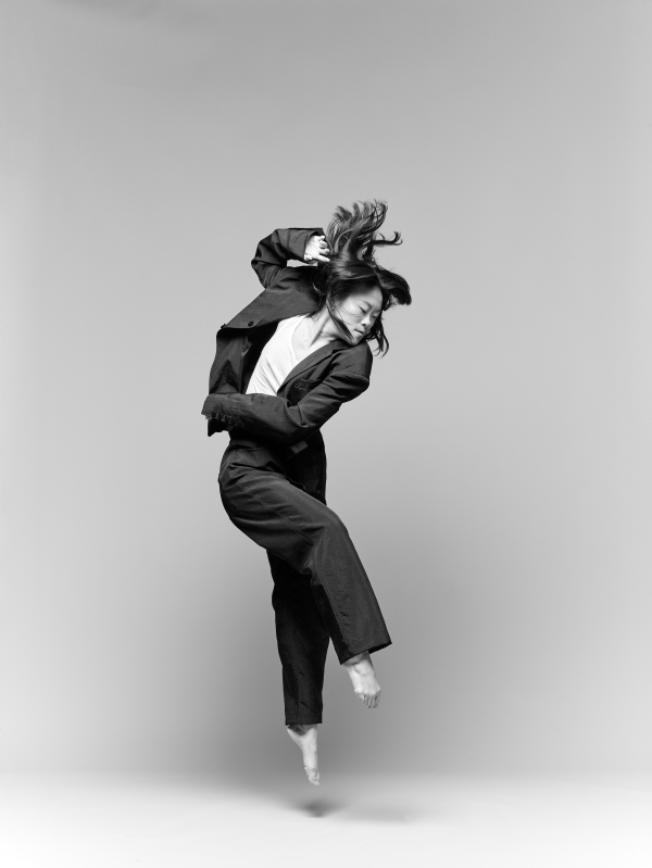 A single dancer in a dark jacket hovering over the floor against a gray background