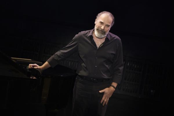 Mandy Patinkin with one arm resting against a piano and smiling.