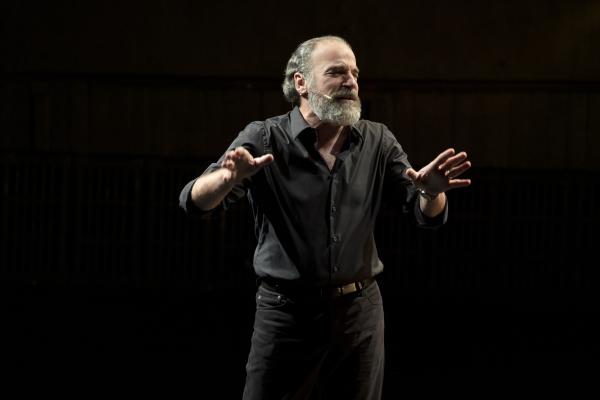 Mandy Patinkin wearing black singing on a dark stage with his hands held forward