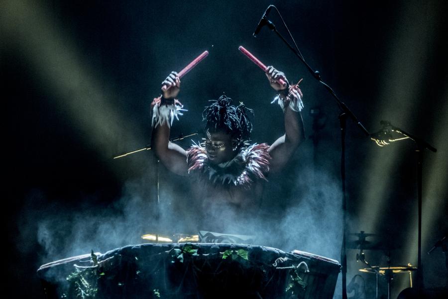 A drummer with arms raised up about to strike a large drum, all dramatically lit