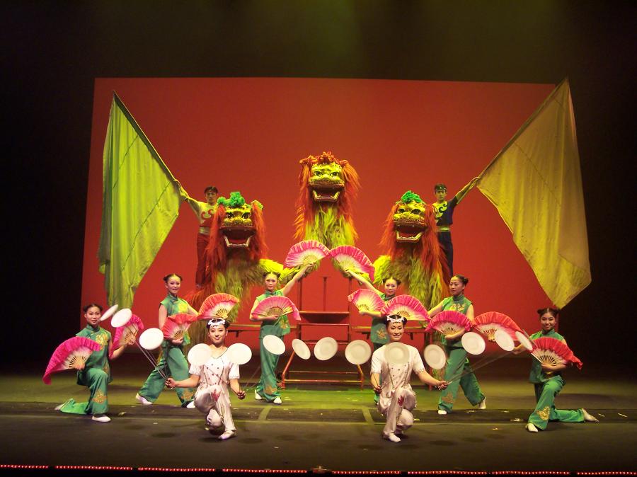 A vibrant stage performance featuring traditional Chinese lion dancers in colorful red and yellow costumes, flanked by performers holding large green flags. In front, dancers in green and white costumes form a semi-circle, skillfully spinning plates and displaying pink fans, all set against a glowing red backdrop under dramatic lighting.