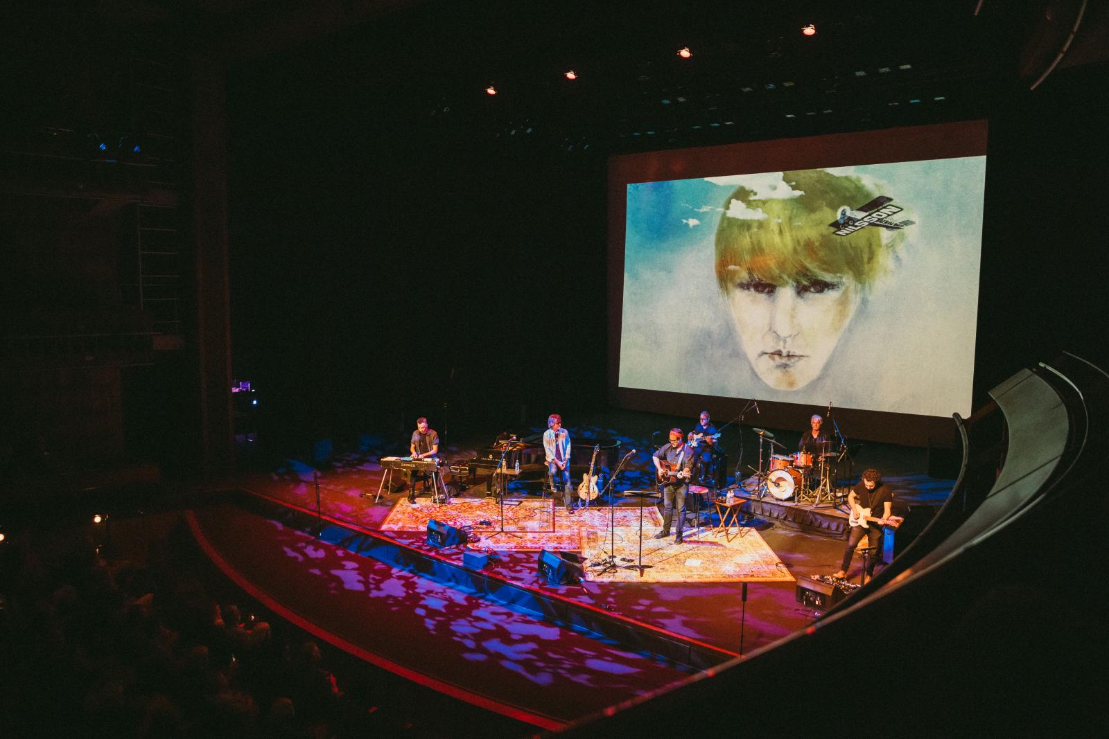 "Live from Laurel Canyon" band performing on stage with instruments under colorful stage lighting, with a large projected image of a painted face and vintage airplane in the background.