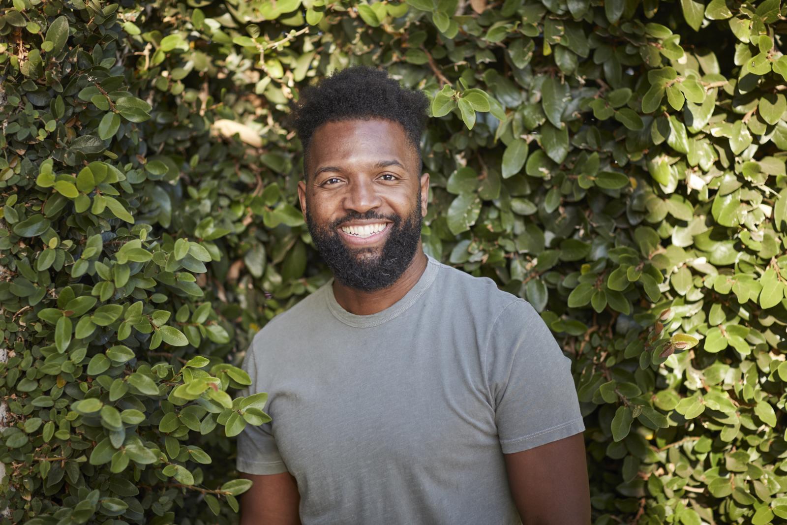 Baratunde Thurston in a gray T-shirt smiling in front of a green hedge