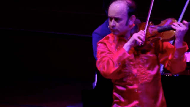Igudesman on the violin with Joo behind him on the piano, on stage, during a performance. Igudesman dances as he plays.